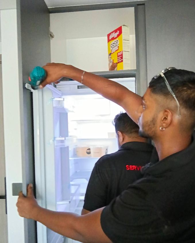 Two technicians adjusting a built-in refrigerator door hinge with a power drill